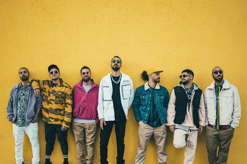 Siete hombres vestidos con ropa casual se apoyan en una pared amarilla.