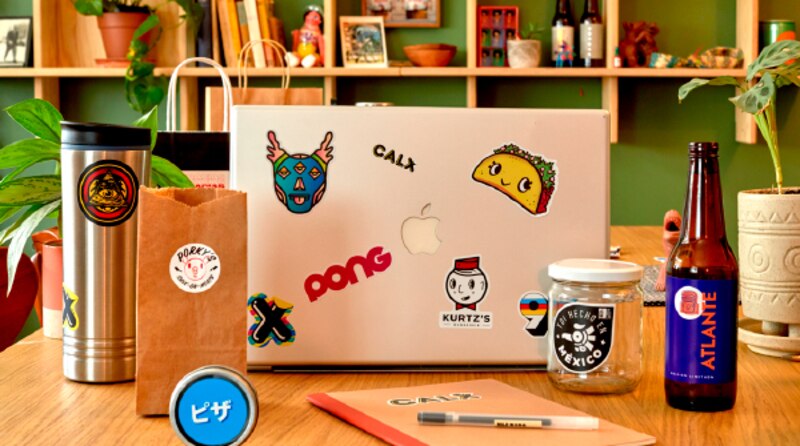Una mesa de trabajo con una computadora portátil, una taza, una bolsa de papel, una cerveza y una planta.