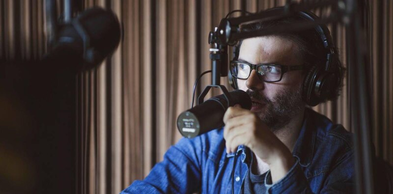 Un hombre joven con barba y gafas está grabando un podcast en un estudio.