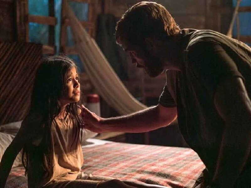 La imagen muestra a un hombre y una niña en una habitación oscura. El hombre está sentado en la cama y la niña está de pie junto a él. El hombre tiene la mano en el hombro de la niña y la está mirando con expresión seria. La niña está mirando al hombre con expresión asustada. La habitación está iluminada por una luz tenue y se pueden ver algunas sombras en la pared.