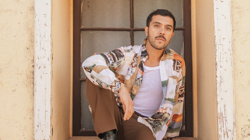 Un hombre joven con barba y camisa estampada se sienta en el suelo junto a una ventana.