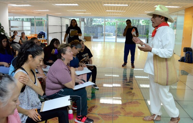 Un hombre con sombrero da una presentación a un grupo de mujeres sentadas en el suelo.