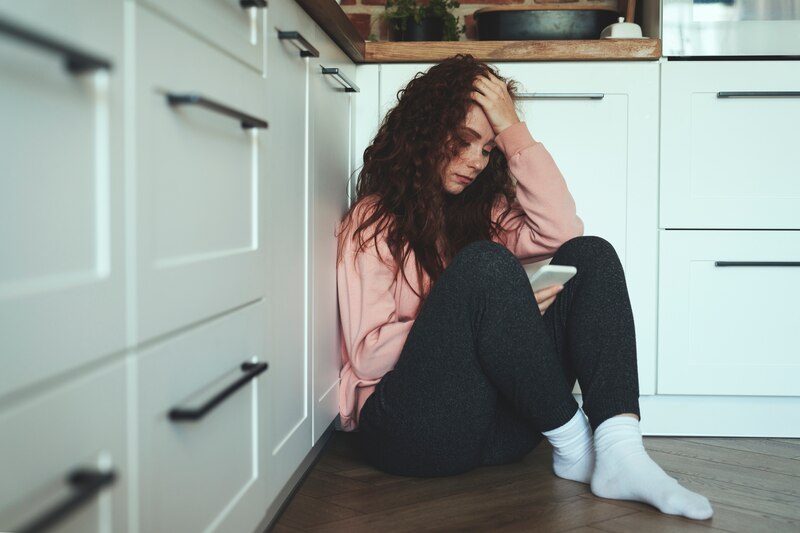 Mujer joven sentada en el suelo de la cocina, con la cabeza entre las manos y mirando su teléfono.