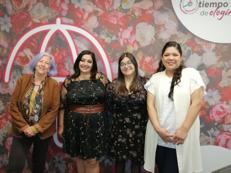 Cuatro mujeres sonríen frente a una pared con un diseño floral