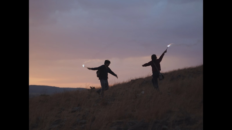 Dos jóvenes corren por un campo al atardecer con bengalas en las manos