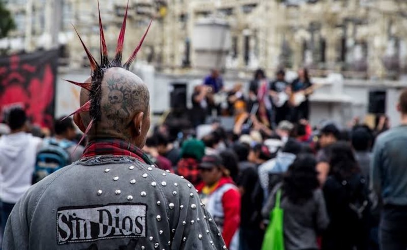 Un hombre con una cresta roja en la cabeza y una chaqueta negra con tachuelas en la espalda está de espaldas en un concierto.