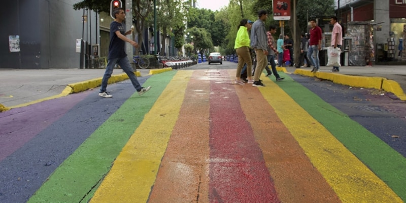 Peatones cruzan una calle con un paso de cebra pintado con los colores del arco iris.