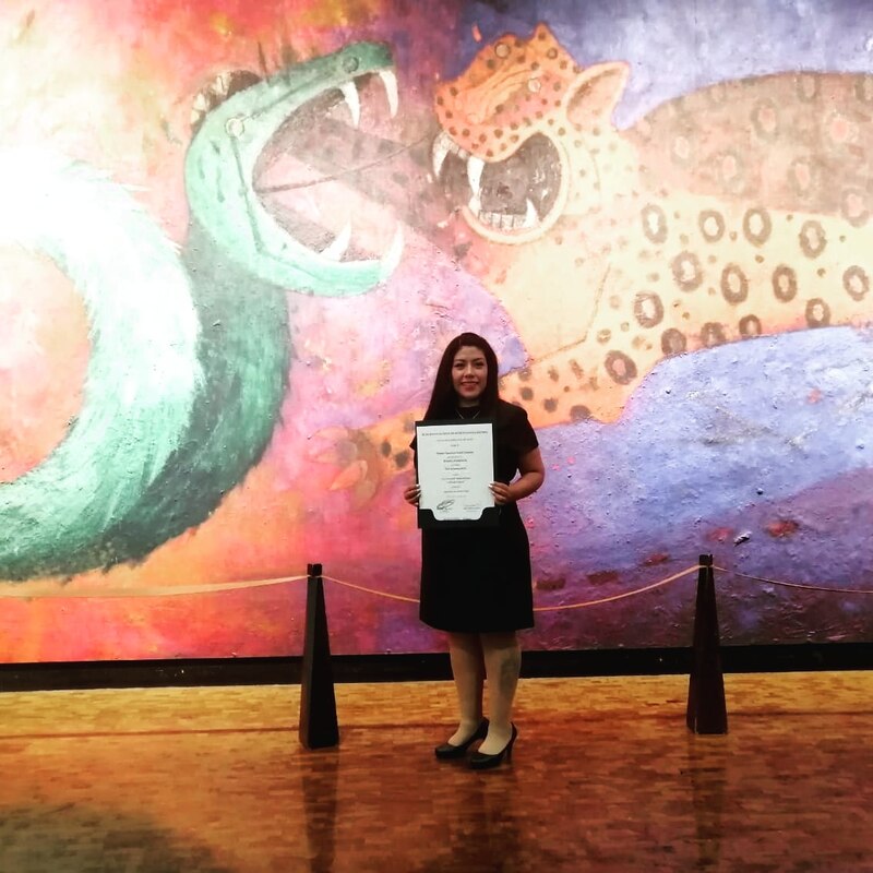 Mujer joven con un certificado en frente de un mural de Rufino Tamayo