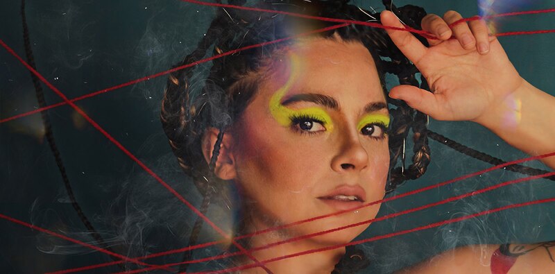 Mujer joven con el pelo largo y negro, maquillaje colorido y brillante, y un vestido negro con hilos rojos que la rodean.