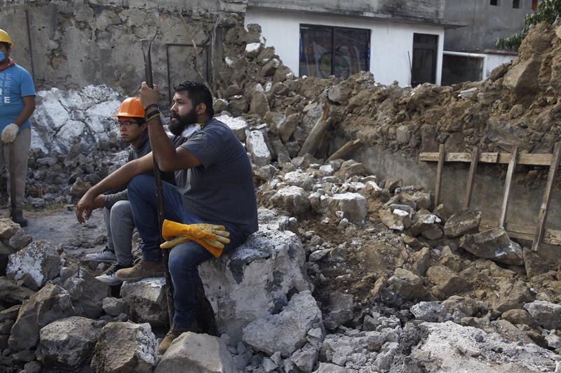 Un hombre sentado en una piedra mira pensativo los escombros de un edificio derrumbado.