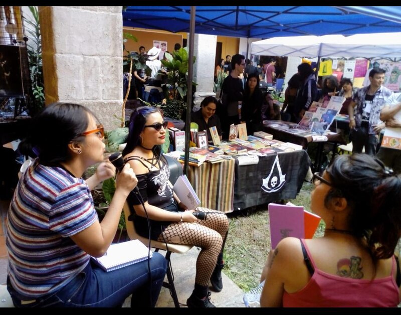 Dos mujeres jóvenes están sentadas en sillas en un mercado al aire libre, una de ellas con una grabadora en la mano.