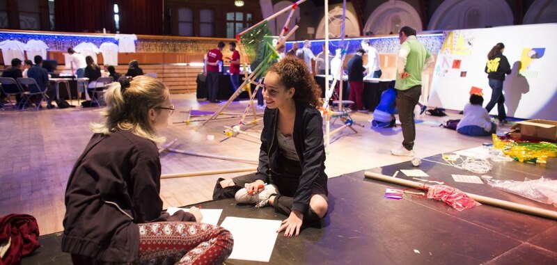 Dos mujeres jóvenes están sentadas en el suelo en medio de una sala grande. Están rodeadas de materiales de arte y parecen estar trabajando en un proyecto creativo.