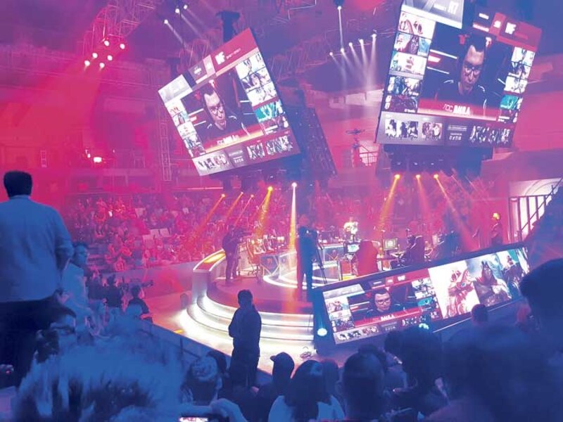 La imagen muestra a un grupo de personas jugando videojuegos en un escenario mientras el público los observa.