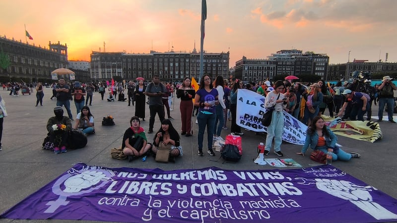Fotografía de una manifestación feminista en la Plaza de la Constitución (Zócalo) de la Ciudad de México al atardecer. La imagen está centrada en un grupo de mujeres sentadas y de pie en la plancha, con una gran lona morada desplegada en primer plano. El mensaje de la lona dice: "Nos queremos Vivas, Libres y Combativas contra la violencia machista y el capitalismo." Al fondo se aprecia el Palacio Nacional y otros edificios históricos del Centro Histórico. Imagen clave de la protesta social y el movimiento feminista mexicano en el Zócalo.