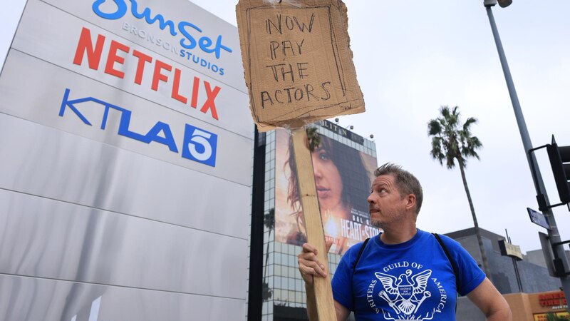 Un manifestante sostiene un cartel frente a un edificio de Netflix que dice "Ahora paguen a los actores".