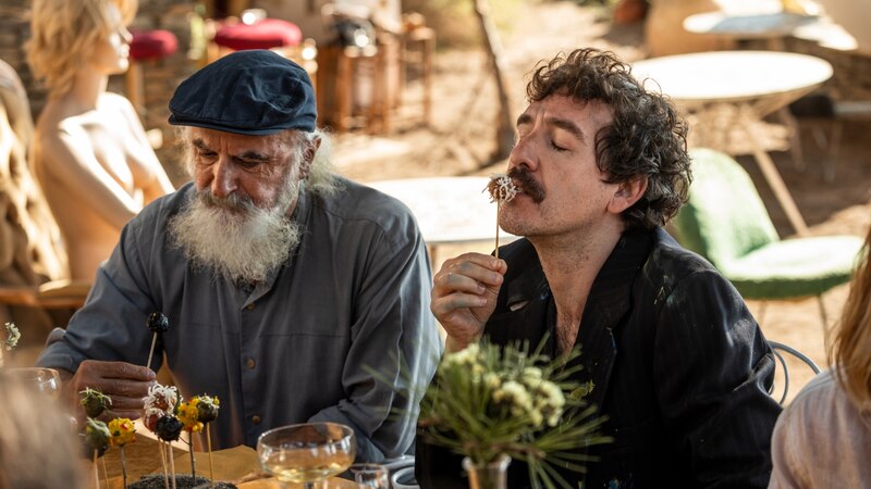 Dos hombres, uno mayor y otro más joven, están sentados a una mesa en el exterior. El hombre mayor tiene barba blanca y lleva una gorra azul. El hombre más joven tiene bigote y el pelo largo y rizado. Ambos están comiendo con los dedos de unos palitos.