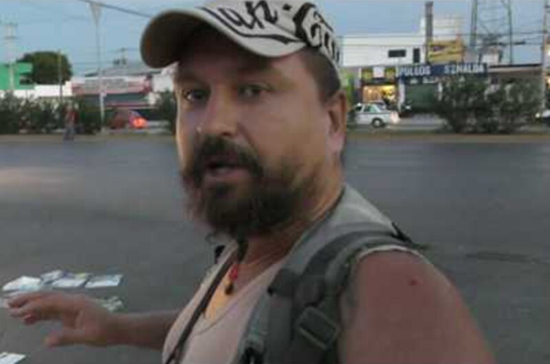 Un hombre barbudo con una gorra negra y una camiseta gris habla a la cámara mientras sostiene un cartel que dice "Se buscan personas para trabajo de limpieza".