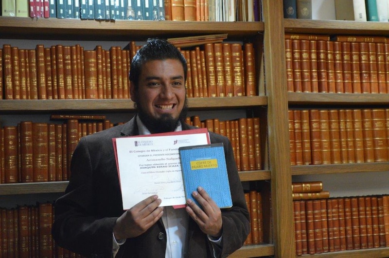 Un joven sostiene un libro y un diploma en una biblioteca.