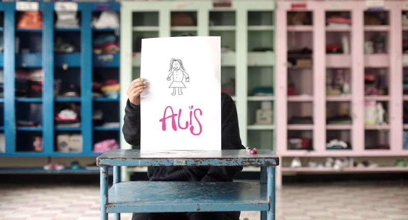 La alumna sostiene un dibujo con la palabra "AUS"