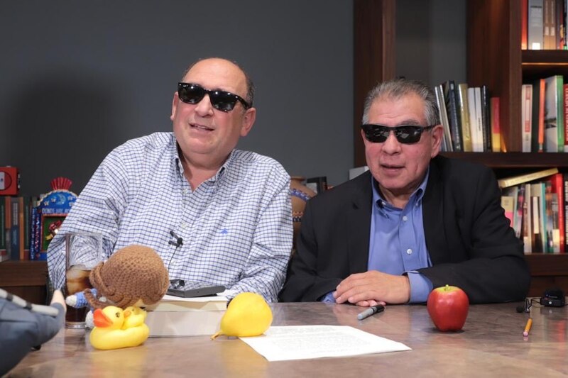 Dos hombres con gafas de sol están sentados en una mesa con un mantel marrón.