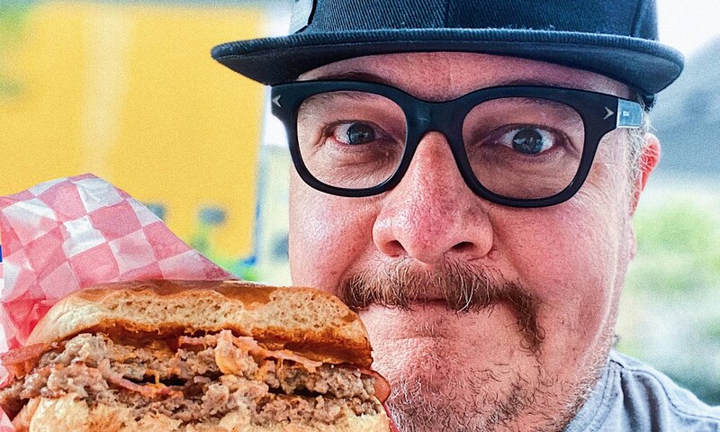 Un hombre con gafas y gorra negra come una hamburguesa con bacon.