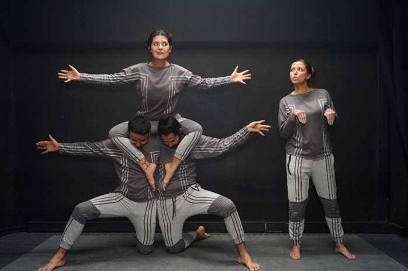 Tres personas vestidas con ropa gris y negra realizan una acrobacia en la que una mujer se encuentra parada sobre los hombros de un hombre y otra mujer se encuentra parada frente a ellos.