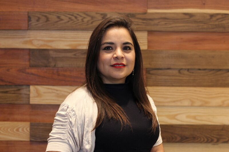 Mujer joven con cabello castaño largo y labios rojos sonríe frente a una pared de madera.