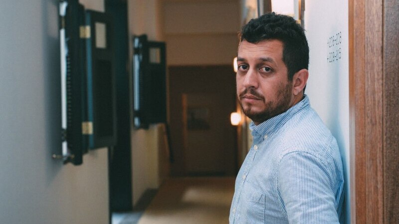Un hombre con barba y camisa azul se apoya en una pared