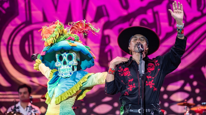 Un cantante con un traje negro y un sombrero rojo actúa en un escenario con un bailarín con un traje de calavera.