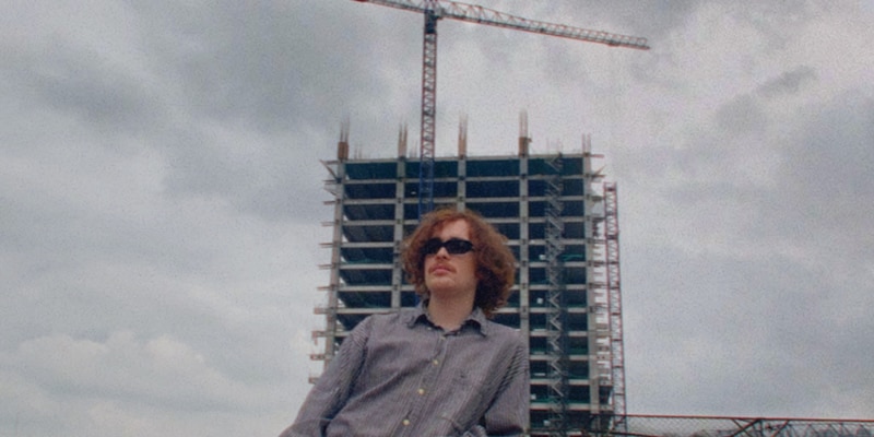 Un joven con gafas de sol y camisa a cuadros se encuentra en la azotea de un edificio en construcción.