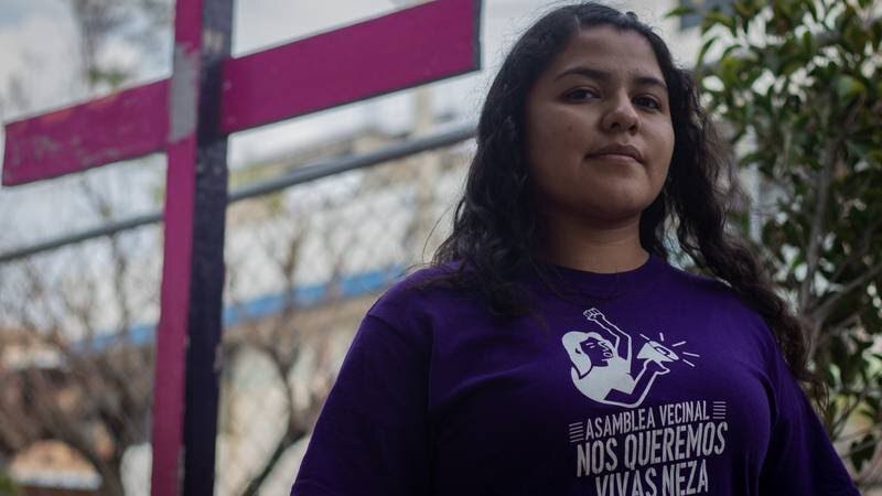 Mujer joven con una camiseta morada con un símbolo feminista y un pañuelo verde en el pelo, de pie frente a una cruz.