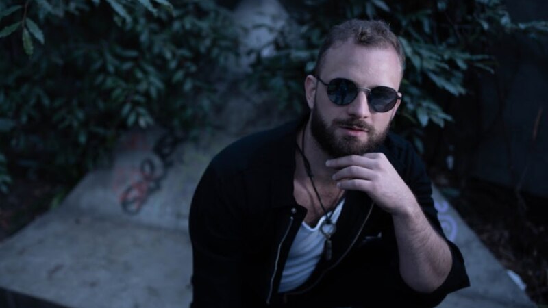 Un hombre con barba y gafas de sol se toca la barba y mira a la cámara.