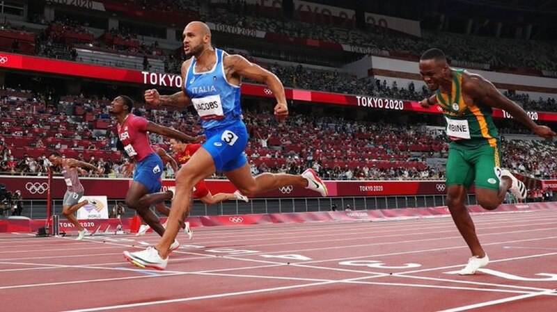 La imagen muestra a tres atletas en una carrera de atletismo. El atleta de la izquierda lleva una camiseta azul y blanca, el del medio lleva una camiseta verde y amarilla, y el de la derecha lleva una camiseta roja y blanca. El atleta de la izquierd