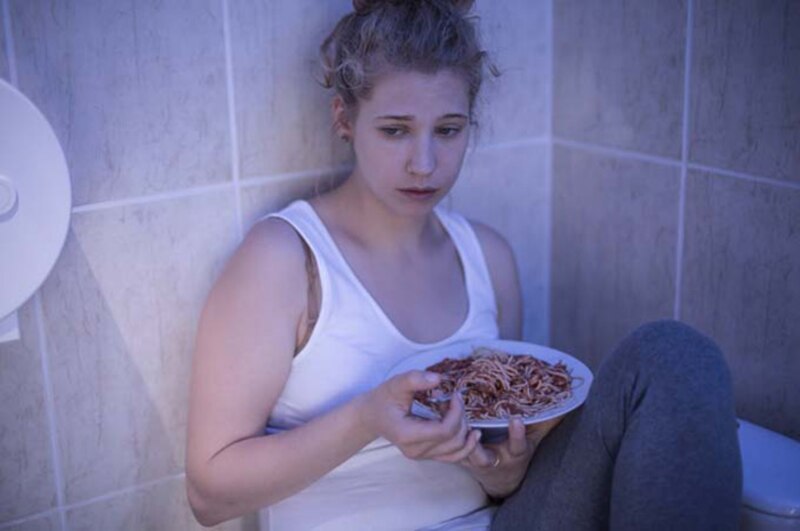 Mujer joven sentada en el suelo del baño con un plato de espaguetis.