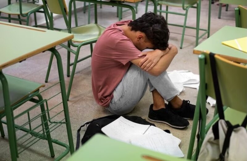 Un estudiante estresado se sienta en el suelo de un aula rodeada de sillas y mesas vacías.