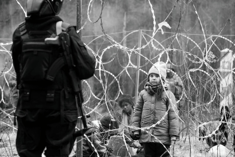 La imagen muestra a una niña mirando a un policía antidisturbios que está de espaldas a ella. La niña está de pie junto a una valla de alambre de espino. La imagen está en blanco y negro.