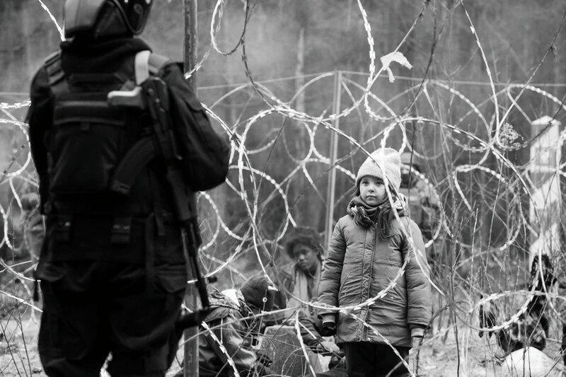 La imagen muestra a una niña mirando a un policía antidisturbios que está de espaldas a ella. La niña está de pie junto a una valla de alambre de espino. La imagen está en blanco y negro.
