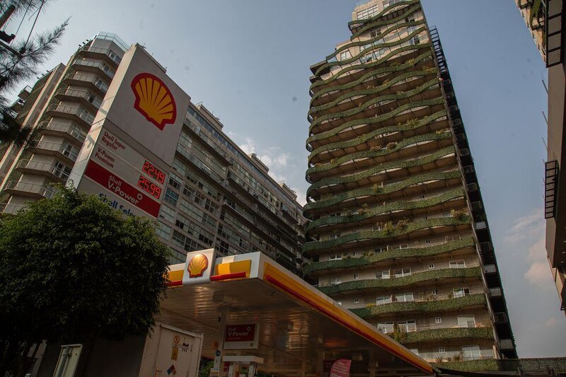 Una gasolinera Shell en primer plano con un edificio alto y verde al fondo