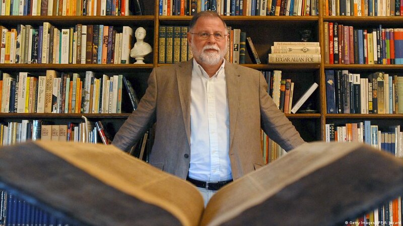 Un hombre sostiene un libro abierto mientras está de pie frente a una estantería llena de libros.