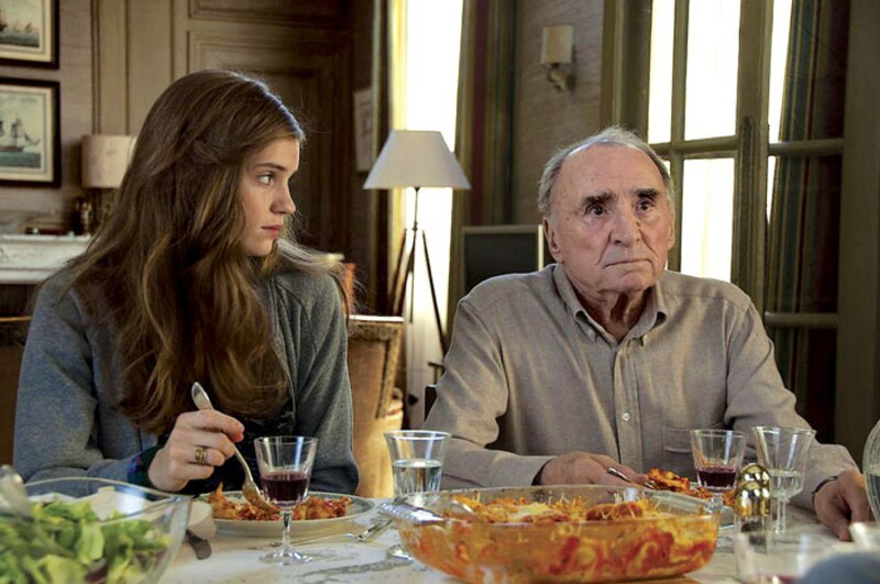 Una joven y un anciano están sentados a la mesa, frente a frente. La joven mira al anciano con expresión seria. El anciano está cabizbajo. Sobre la mesa hay una fuente de pasta y dos copas de vino tinto.