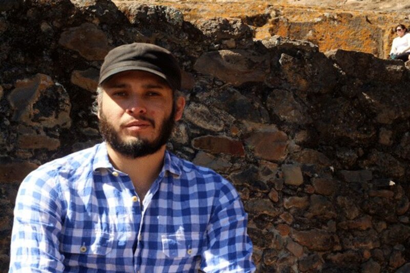 Retrato de un hombre barbudo con camisa a cuadros azules y gorra negra sentado junto a un muro de piedra.