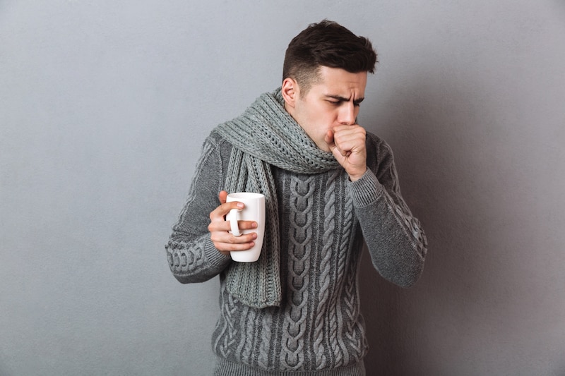 Hombre enfermo tosiendo sosteniendo una taza de té sobre una pared gris de fondo