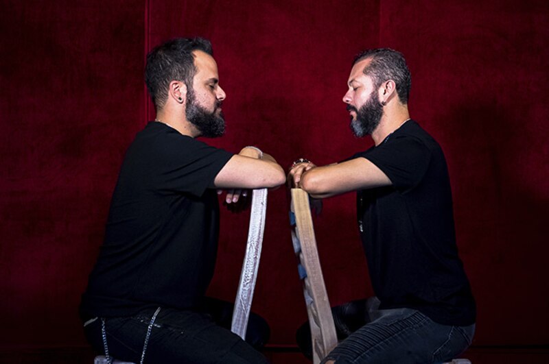 Dos hombres idénticos con barba se sientan uno frente al otro en sillas separadas por una pared roja.