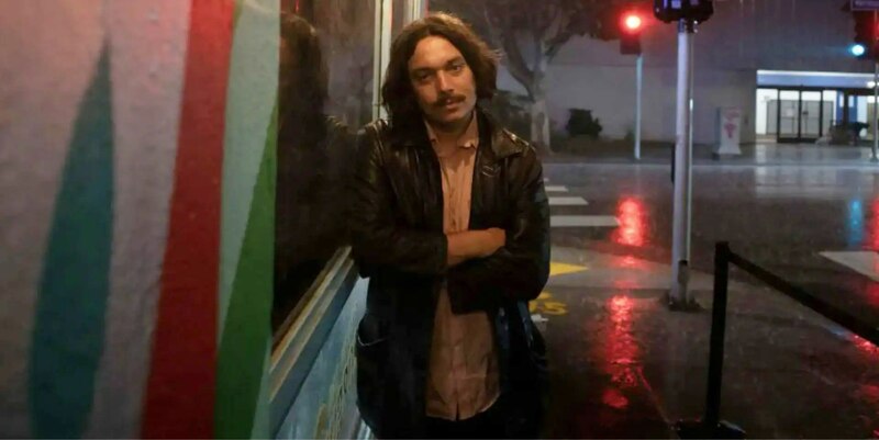 Un hombre se encuentra en una calle mojada por la lluvia, con las luces de la ciudad reflejándose en el agua.