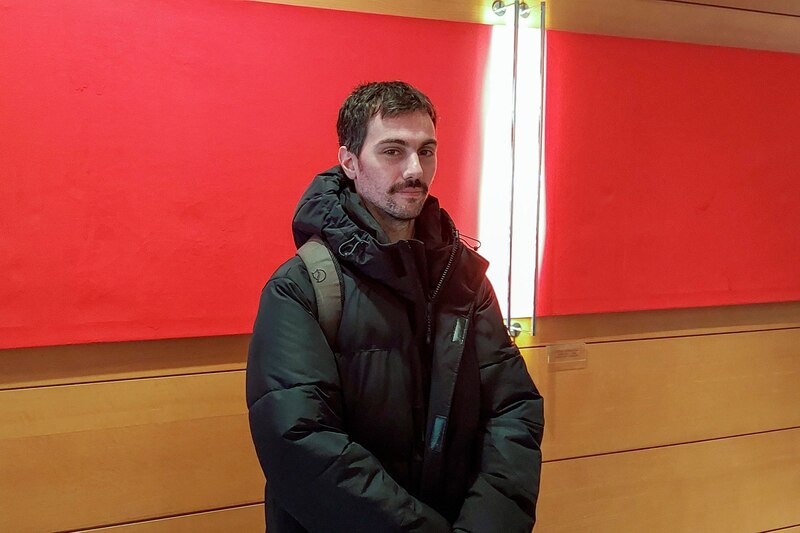 Un hombre joven con barba y el pelo corto y negro, vestido con una chaqueta negra y una mochila, se encuentra de pie frente a una pared roja.