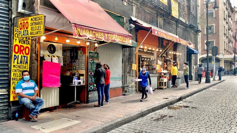 La imagen muestra a un hombre sentado en la banqueta, frente a una tienda de compra y venta de oro y plata. En la tienda hay dos personas más, una está sentada y otra de pie. En la calle hay varias personas caminando.