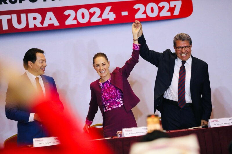 La imagen muestra a tres personas en un podio. La persona en el centro es una mujer, y las otras dos son hombres. La mujer está vestida con un traje rosa y negro, y los hombres visten trajes oscuros. La mujer está sonriendo y levantando la mano derecha, y los hombres también están sonriendo. En el fondo hay un letrero que dice "Morena 2024-2027".