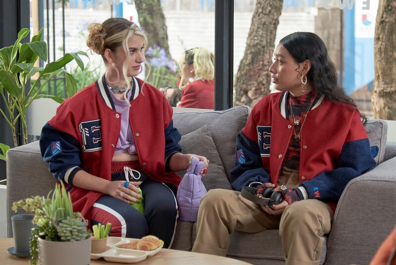 Dos mujeres jóvenes están sentadas en un sofá, una de ellas con una chaqueta roja y la otra con un chaleco negro.