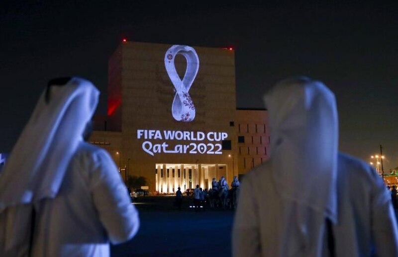 La imagen muestra a dos hombres mirando un edificio con el logo de la Copa Mundial de la FIFA 2022 proyectado en él.