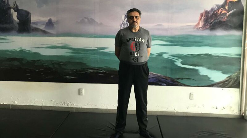 Un hombre de mediana edad con una camiseta gris y pantalones negros está de pie en una colchoneta de yoga en el interior de un gimnasio.
